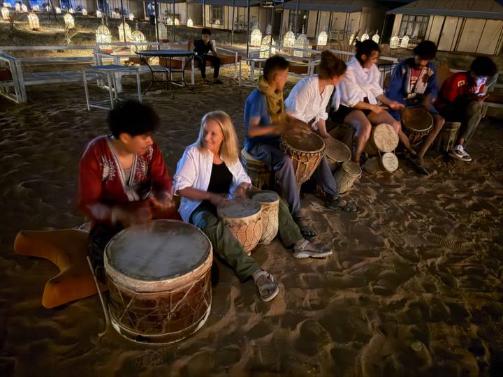 Nighttime drum circle on desert sand with motion-blurred participants playing hand drums.