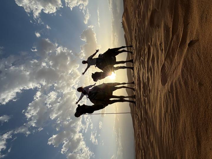Silhouetted riders on camels strike joyful poses against a desert sunset.