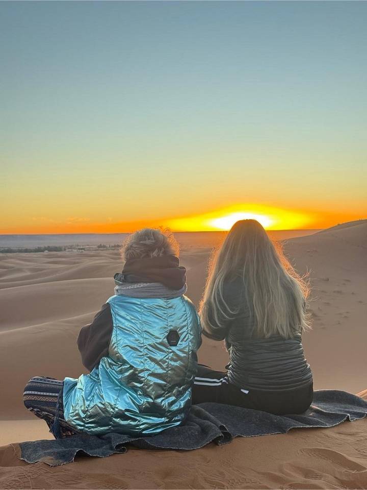Two women sit on a dune watching the sun dip below the Sahara horizon
