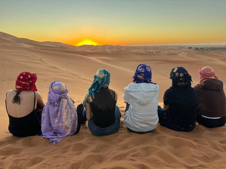 Six friends wearing colorful scarves sit side by side on a dune watching a vivid sunset