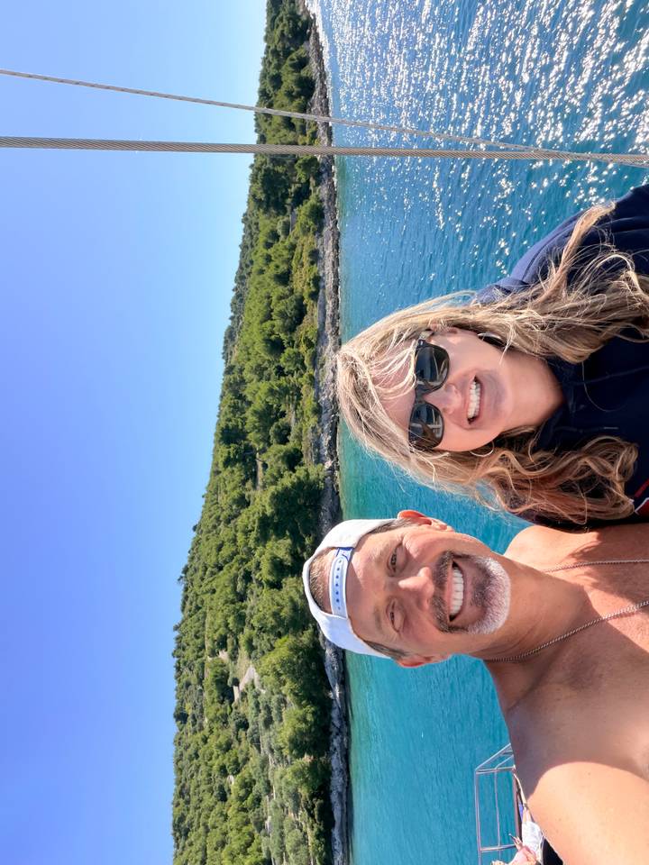 Couple selfie with clear turquoise water and forested coastline under bright blue sky.