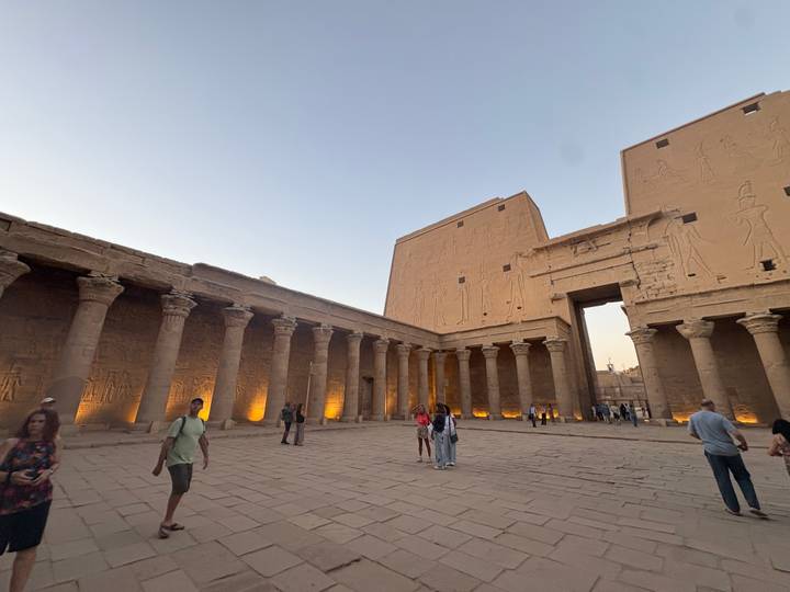 Interior court of Edfu Temple with columns lit by uplights while visitors wander during twilight.