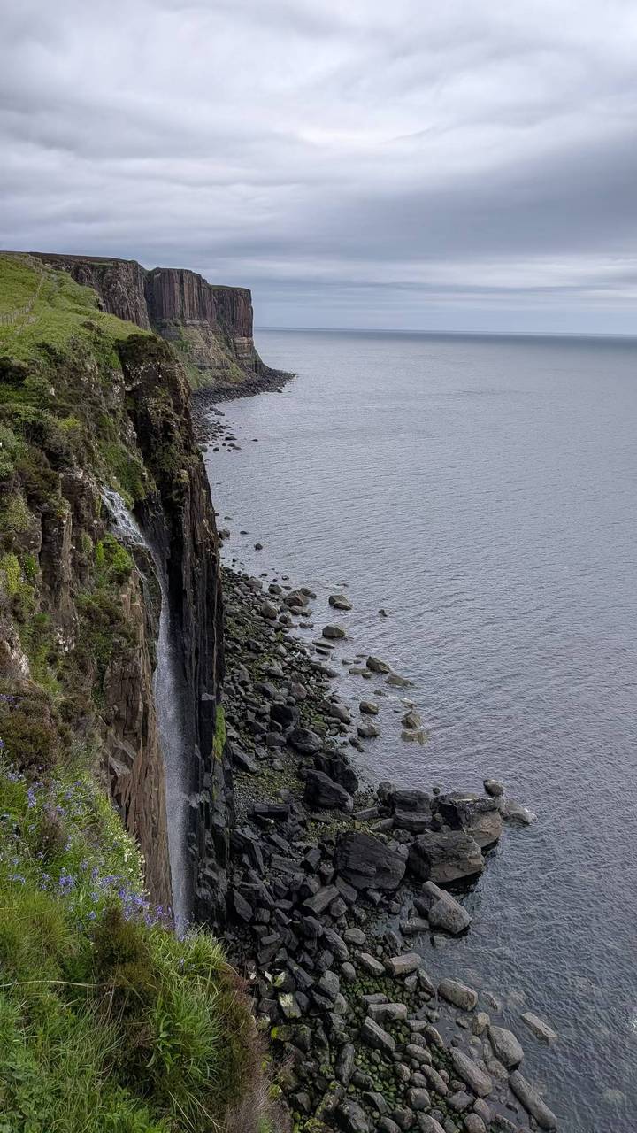 Vertical basalt cliffs with a slender waterfall plunging into rocky shore and sea.