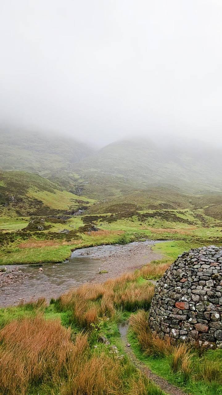 Misty green valley with a stream winding through grassy hills under low clouds.