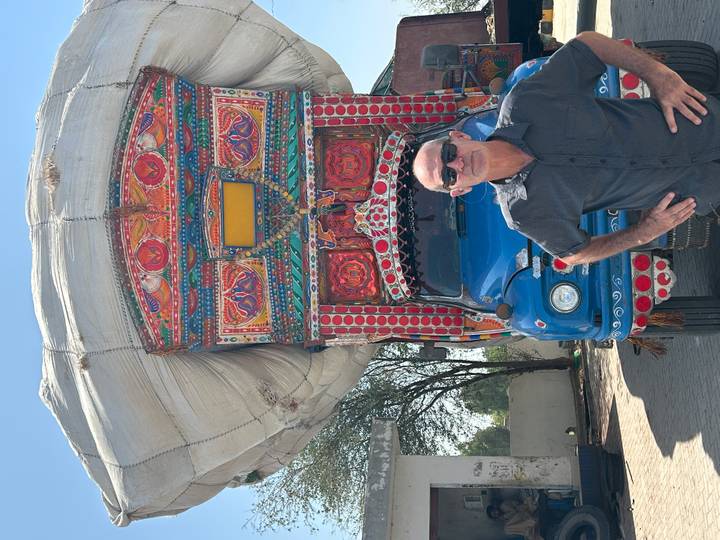 Traveler standing in front of a highly decorated blue Pakistani truck carrying an enormous white tarp bundle on top.