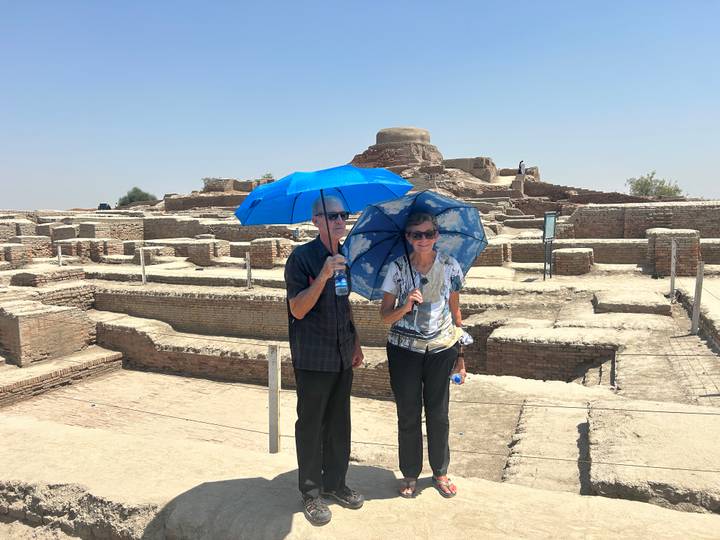 Older couple under bright blue umbrellas exploring sun-baked archaeological ruins of an ancient brick settlement.