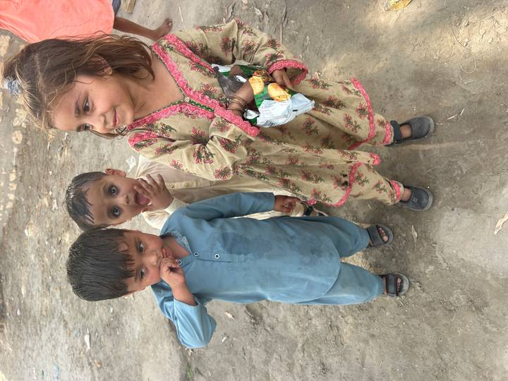 Three small children in traditional clothes standing on dusty ground, one girl holding a packet of snacks.