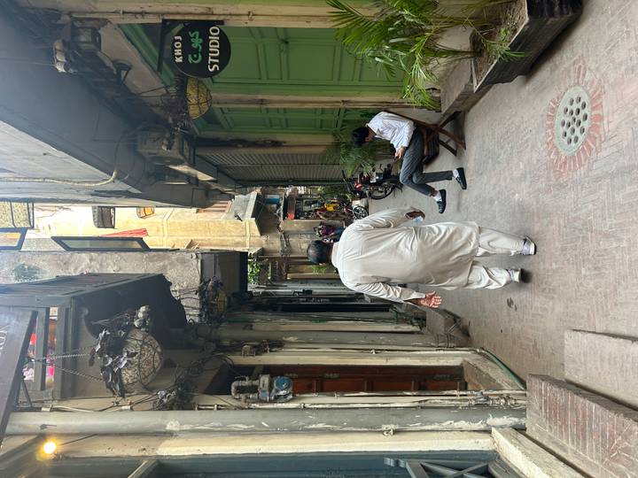Narrow alleyway with hanging lights and balconies where a man in white walks away and another sits on a chair using his phone.