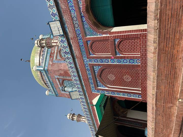 Brick and tile mausoleum with green dome and patterned facade under a bright blue sky with a few birds flying.