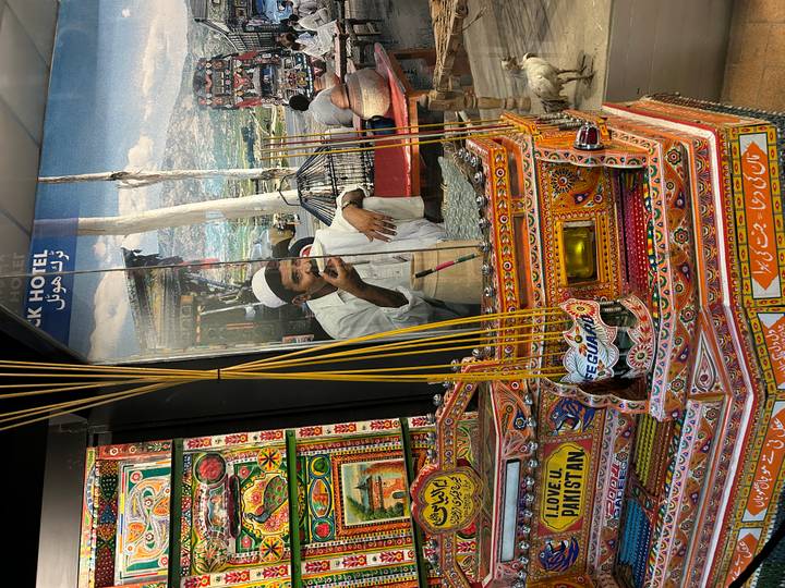 Colorful display of miniature decorated Pakistani trucks and incense sticks with a photo of a man drinking tea in the background.