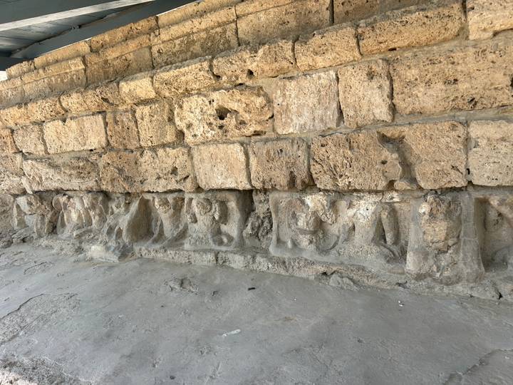 Eroded stone wall base featuring ancient carved reliefs of seated figures inside an archaeological site.