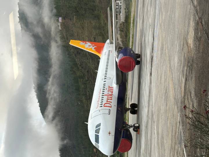 Royal Bhutan Airlines (Drukair) Airbus parked on Paro airport apron with misty forested hills behind.