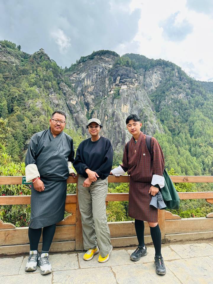 Three travelers in traditional and casual attire stand against a wooden railing with the Tiger's Nest cliff in the background.