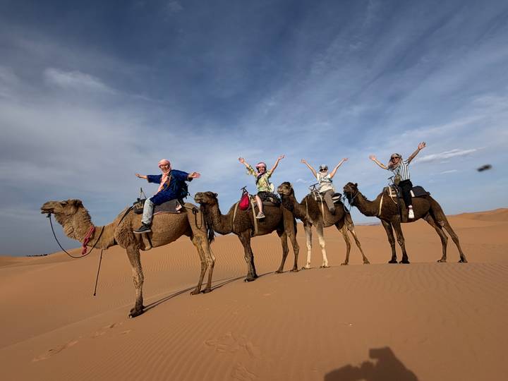 Travellers riding a line of camels across rolling Sahara dunes with arms raised in joy