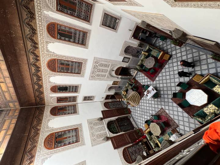 View looking down into an ornate Moroccan riad courtyard with patterned tile floor, low tables, green velvet seating and carved wooden balconies.