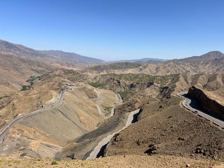 Sweeping aerial view of winding asphalt road snaking through barren brown mountains under a clear blue sky.