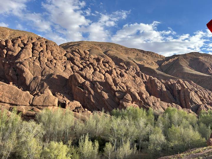 Rugged red rock formations rising against a partly cloudy blue sky with green trees in the foreground.
