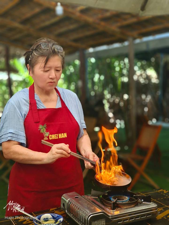 A woman wearing a red apron cooks over an open flame in a rustic outdoor kitchen.