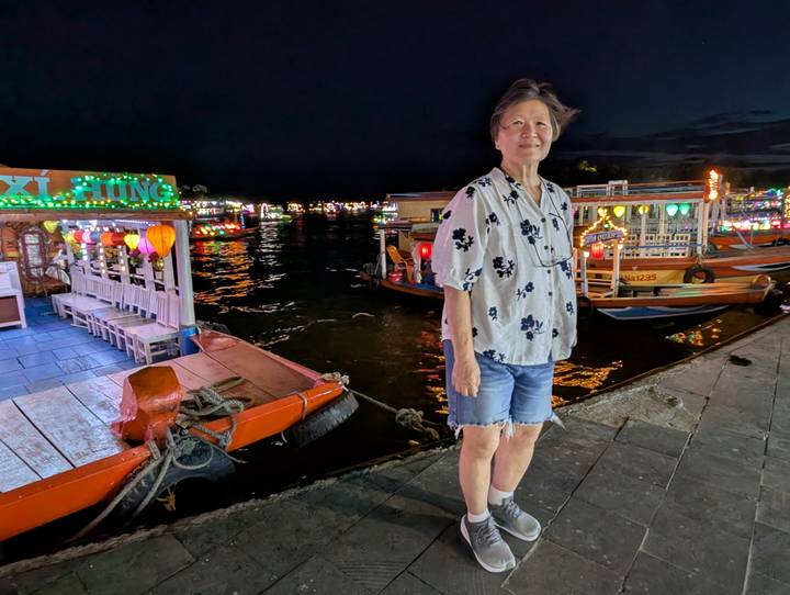 A woman stands on a riverside promenade at night surrounded by colorful lantern-lit boats.