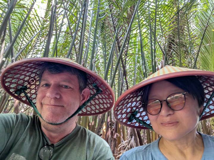 A couple wearing conical hats smile beneath tall palm fronds in lush wetland vegetation.