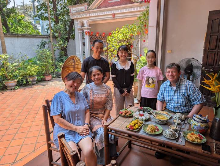 A Vietnamese host family and two guests enjoy a traditional meal in a sunny courtyard.