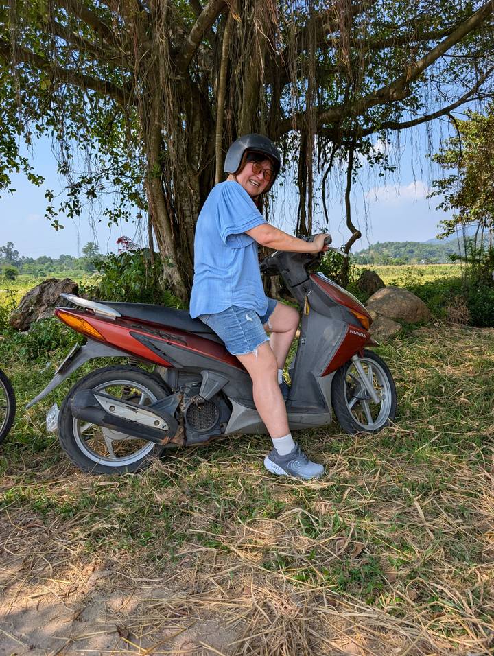 A woman in casual clothes sits astride a red scooter beneath a shady tree in the countryside.