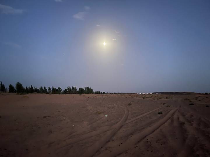 Moonlit Sahara desert landscape with faint camp tents visible on the horizon