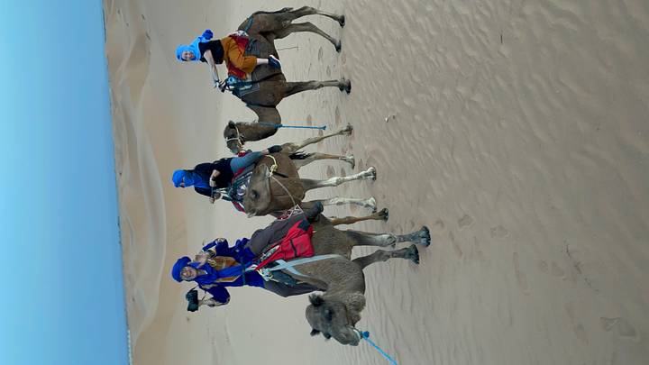 Three travelers dressed in blue headscarves riding camels on sand dunes during daylight