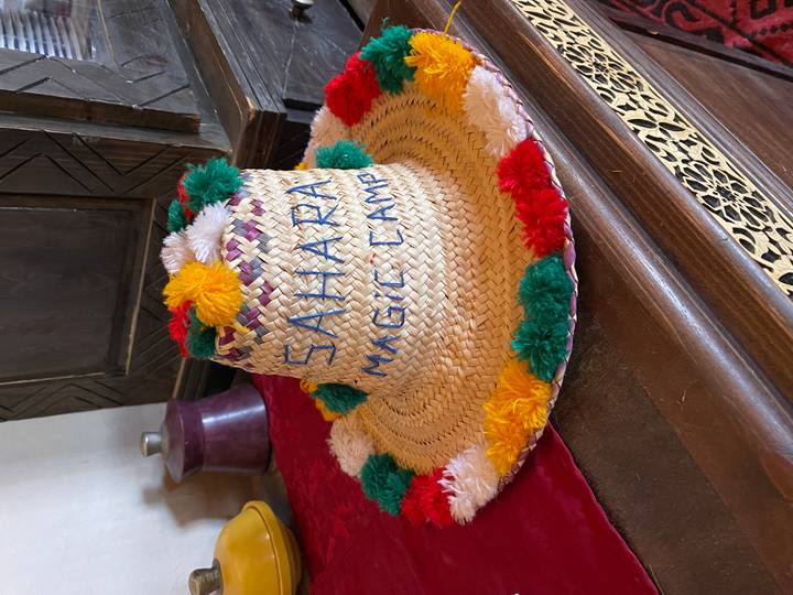 Hand-woven straw hat decorated with multicolored pompoms labeled 'Sahara Magic Camp'