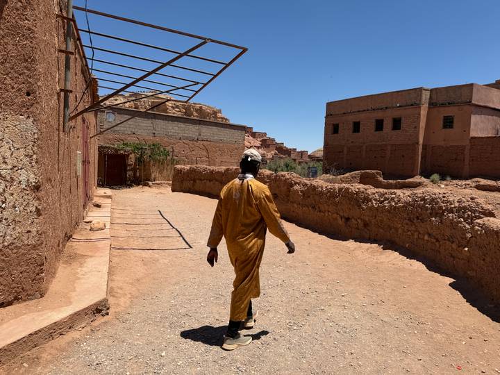 Man in traditional robe walking a dusty lane lined with earthen walls in a desert village.