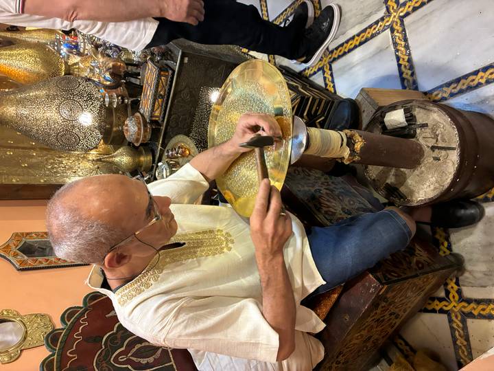 Moroccan artisan meticulously hammering a brass plate in a traditional workshop filled with metal lamps
