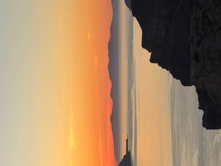 A vivid sunset over calm sea waters with dark silhouettes of distant islands beneath an orange and pink sky.