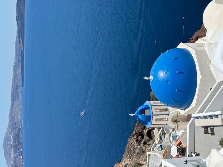 Iconic white church with bright blue dome perched on cliffs above the deep-blue Aegean Sea with a small boat below.