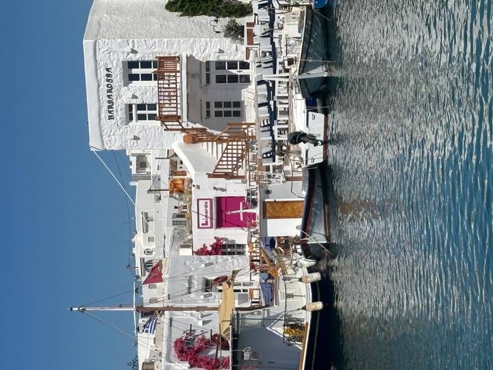 Charming waterfront lined with whitewashed buildings and moored fishing boats under a clear blue sky; a sailor works on a small vessel.