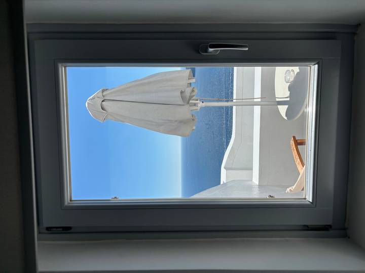 View through a white-framed window to a balcony umbrella and endless blue sea under cloudless sky.