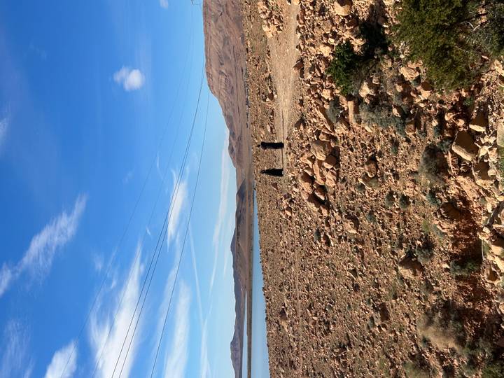 Two distant figures walk along a rocky lakeside plain beneath a bright blue sky.