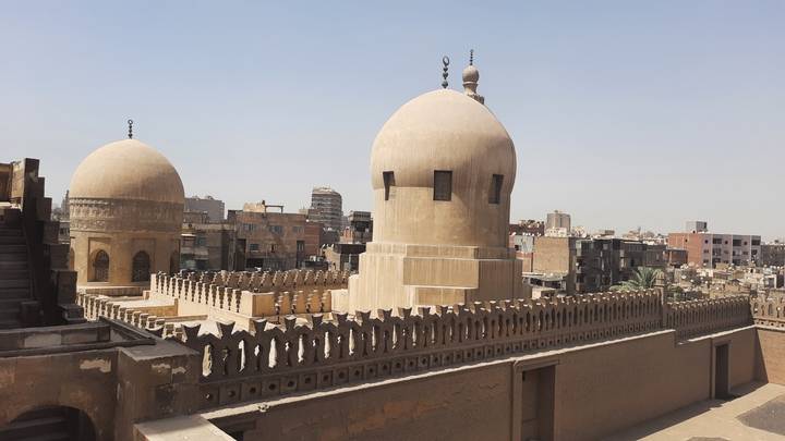 Twin domes and crenellated walls of an old Cairo mosque overlook densely packed city buildings.