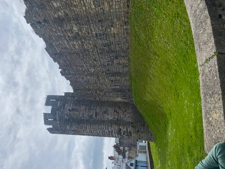 Stone defensive wall and round tower of Caernarfon Castle under grey sky.