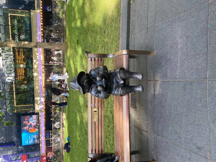 Bronze Paddington Bear statue seated on bench in busy Leicester Square garden.