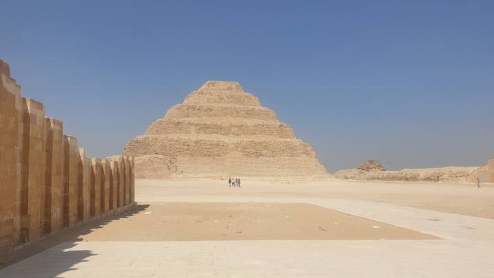 Wide shot of the Step Pyramid of Djoser rising from the sandy desert plateau under a clear blue sky.