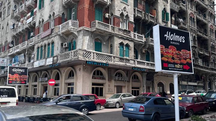 Busy city corner with historic building featuring green shutters, billboard advertising burgers and several parked cars.