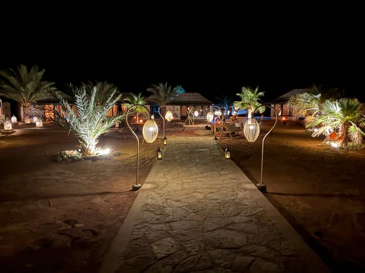Elegant desert camp lit by lanterns and palm trees lining a stone path at night.