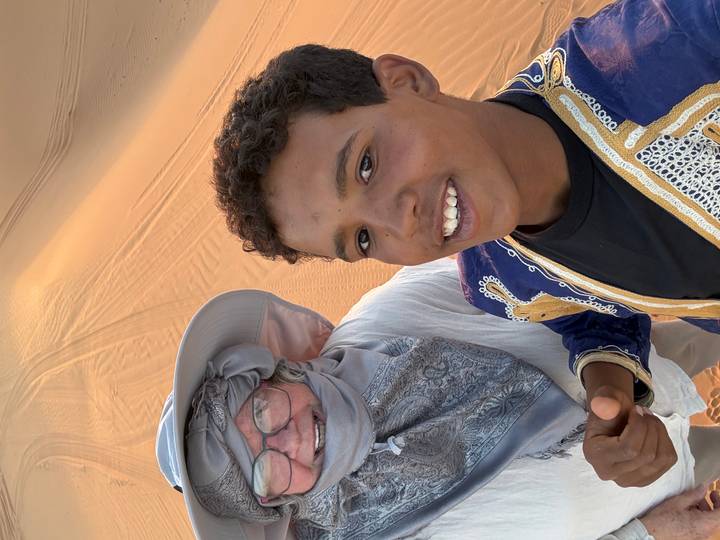 Smiling young local boy and older tourist pose for a selfie amid golden Sahara sand dunes.