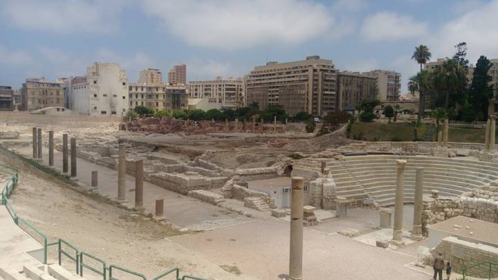 Ruins of a Roman theatre and columns in Alexandria with modern city buildings in the background.