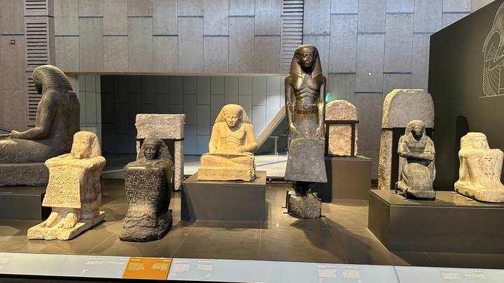 Row of ancient Egyptian statues displayed inside a modern museum gallery with visitors walking by.