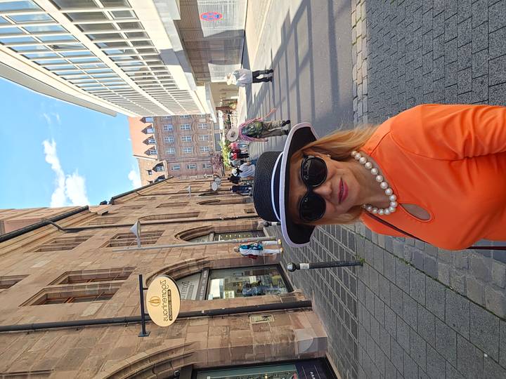 Stylish woman in orange dress and hat poses on a cobblestone pedestrian street in a European old town.