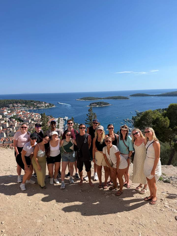 Group of travelers smiling at a cliff-top viewpoint overlooking Hvar town, small islands and deep blue Adriatic waters under a clear sky.