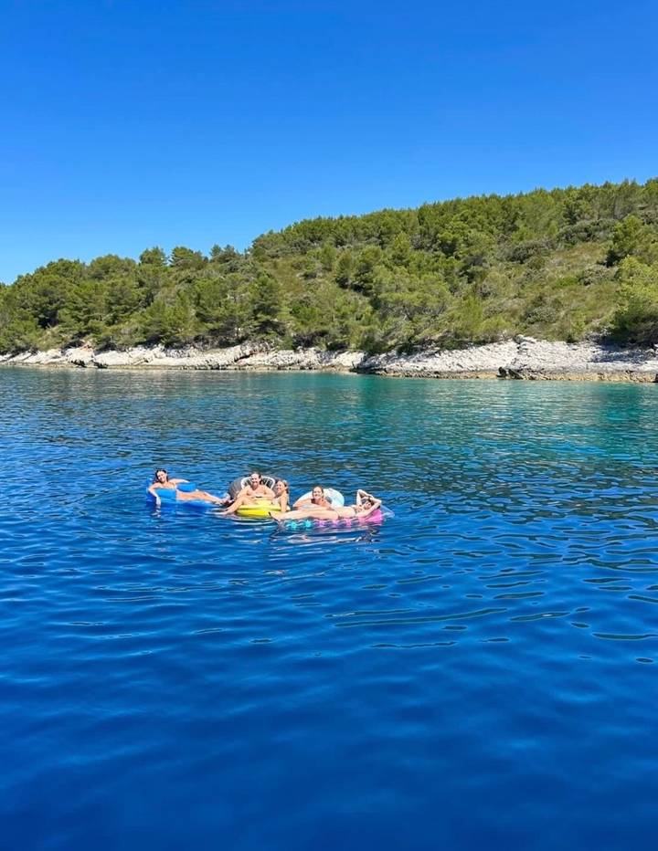 Small group lounging on colorful inflatables in calm, clear blue Adriatic water beside a pine-covered rocky coastline.