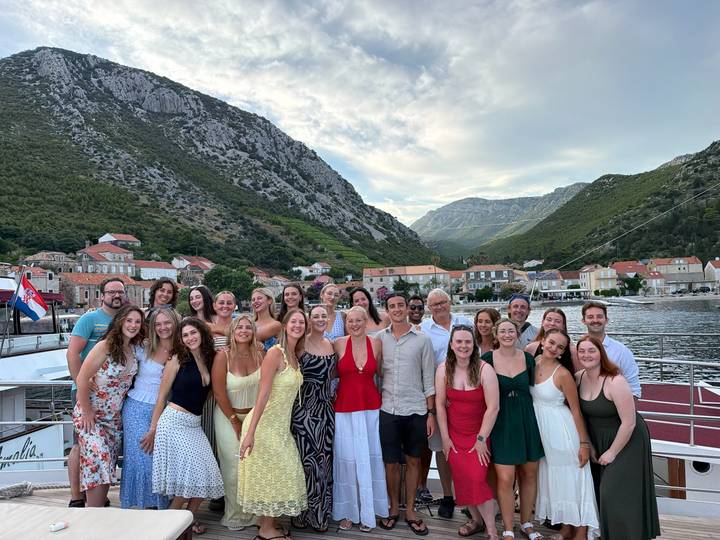 Large tour group posing on a vessel docked in a scenic Croatian harbor framed by steep green mountains and a village.