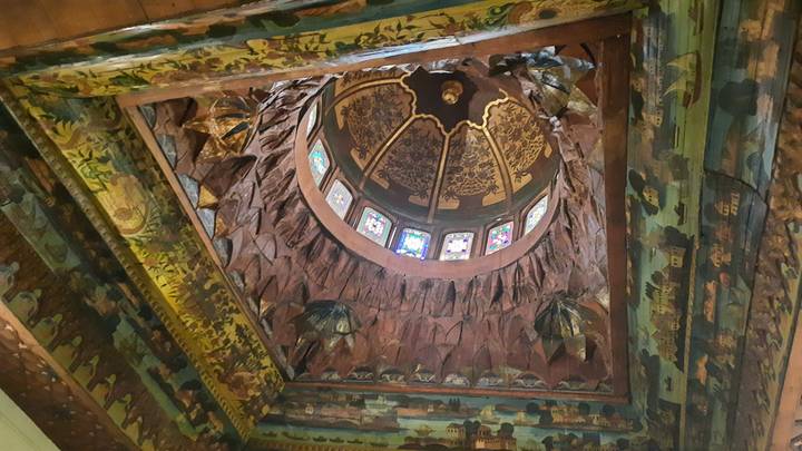 Intricately painted and carved domed ceiling with colorful stained-glass windows viewed from below.